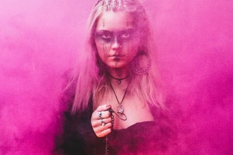 Power Dressing - Blond scary woman with makeup and in dress holding glowing lamp while standing in pink colored smoke looking at camera