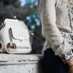 Fashion - woman wearing beige and red floral top leaning on gray concrete slab with white leather bag ontop