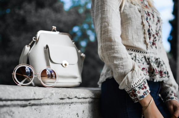 Fashion - woman wearing beige and red floral top leaning on gray concrete slab with white leather bag ontop