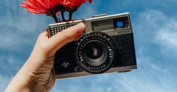 Vintage Revival - From below crop anonymous female photographer holding vintage photo camera with blooms against sky