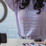 Fashion's Influence - Stylish young African American female influencer in sweater adjusting long dark hair while sitting at wooden table before recording makeup tutorial video on smartphone