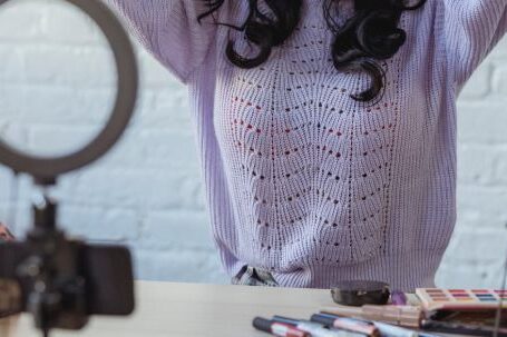 Fashion's Influence - Stylish young African American female influencer in sweater adjusting long dark hair while sitting at wooden table before recording makeup tutorial video on smartphone