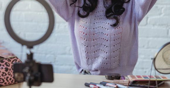 Fashion's Influence - Stylish young African American female influencer in sweater adjusting long dark hair while sitting at wooden table before recording makeup tutorial video on smartphone